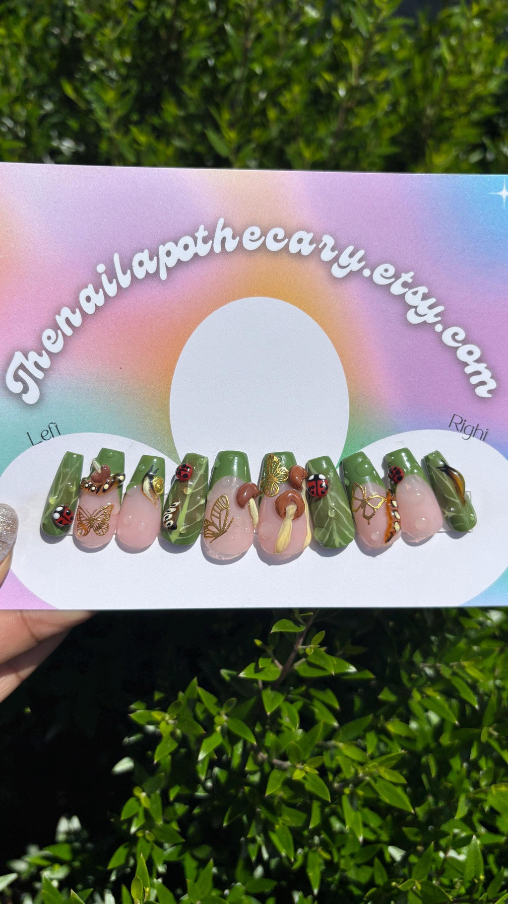 Nail art display with green and pink designs featuring brown capped mushrooms with yellow stems , green French tips with a nude pink base with gold butterfly and 3d lady bugs and snails spread throughout the set on a rainbow-colored card with 'thenailapothecary.etsy.com' text.