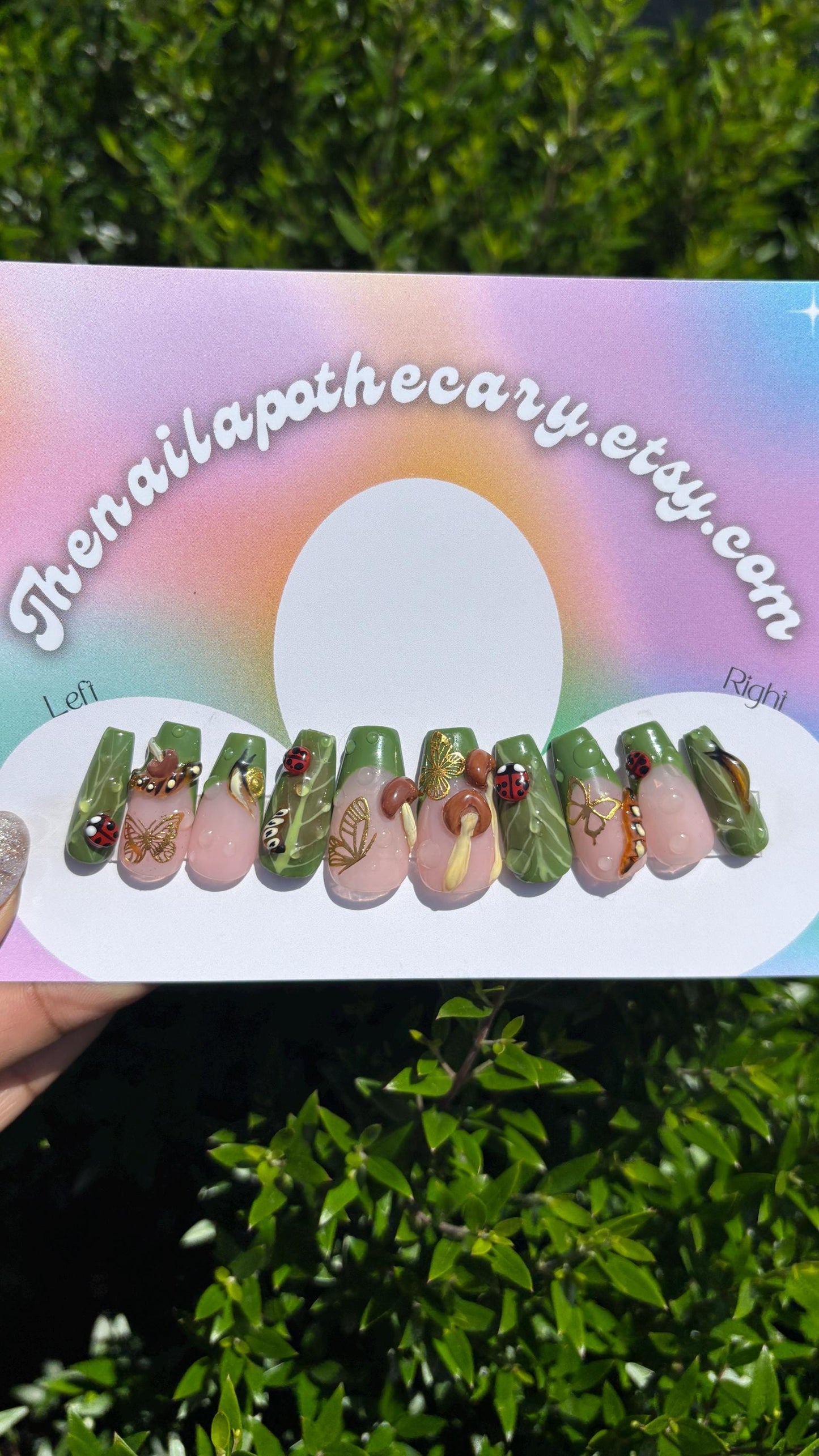 Nail art display with green and pink designs featuring brown capped mushrooms with yellow stems , green French tips with a nude pink base with gold butterfly and 3d lady bugs and snails spread throughout the set on a rainbow-colored card with 'thenailapothecary.etsy.com' text.